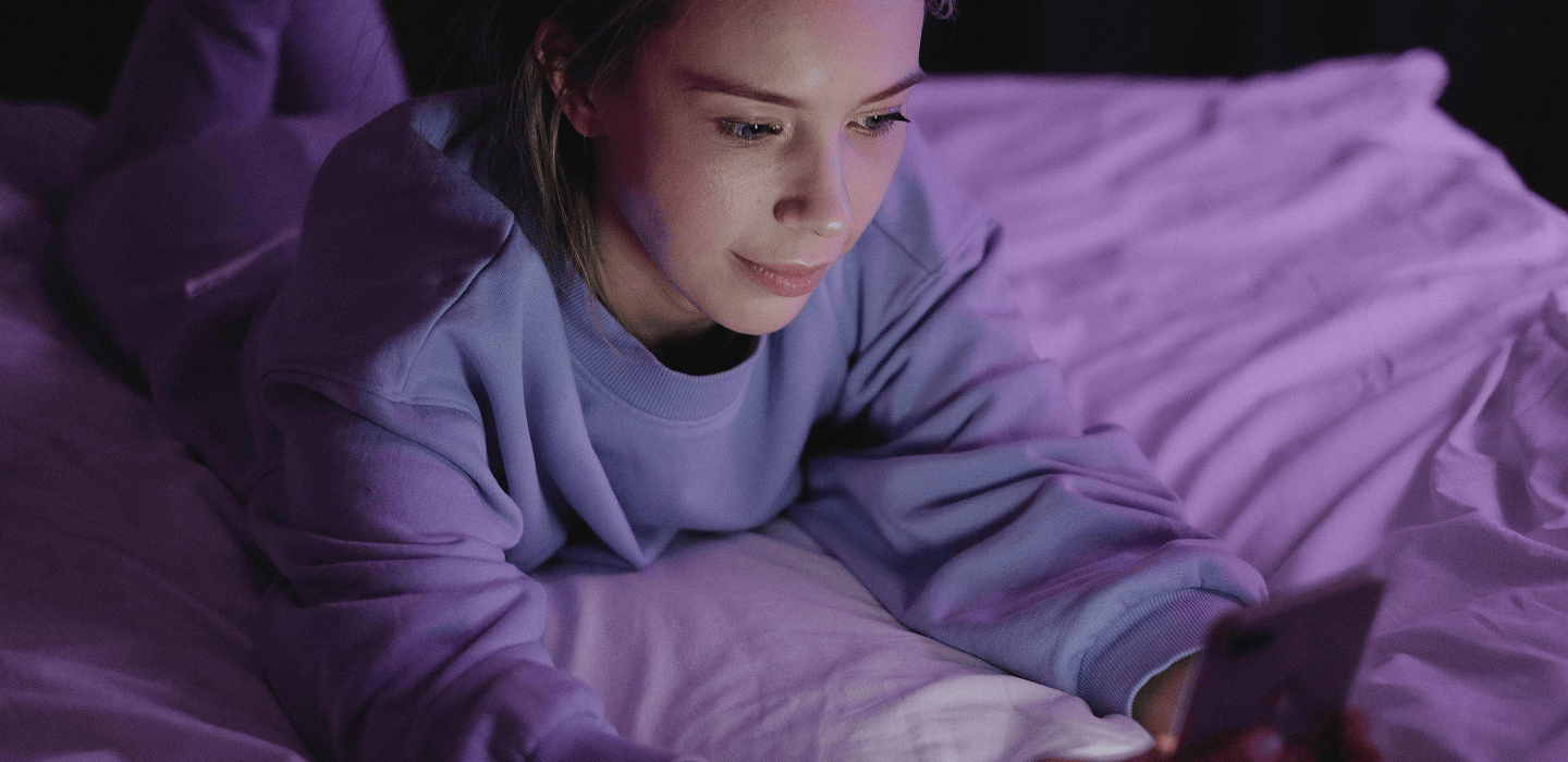 Une personne est allongée sur un lit avec un téléphone intelligent, éclairée par un doux éclairage violet et bleu dans une pièce sombre.