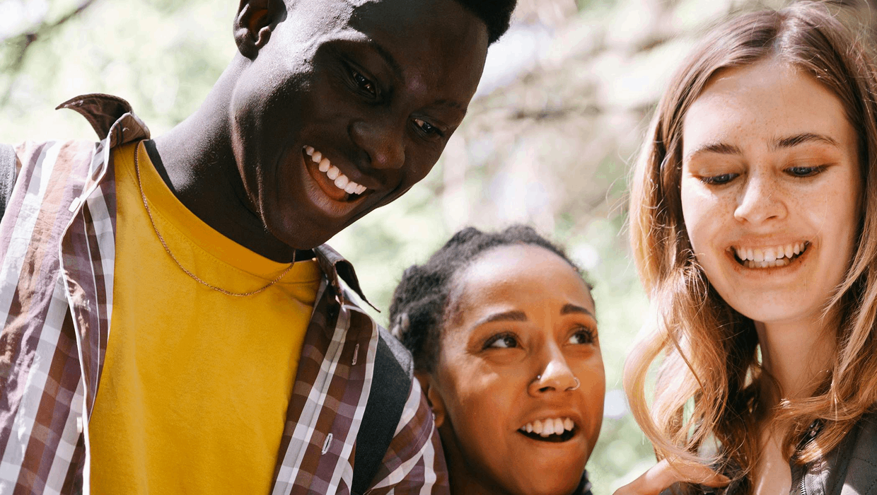 Trois personnes se tiennent l'une près de l'autre à l'extérieur, regardant vers le bas en souriant, avec une lumière naturelle douce et de la verdure à l'arrière-plan.