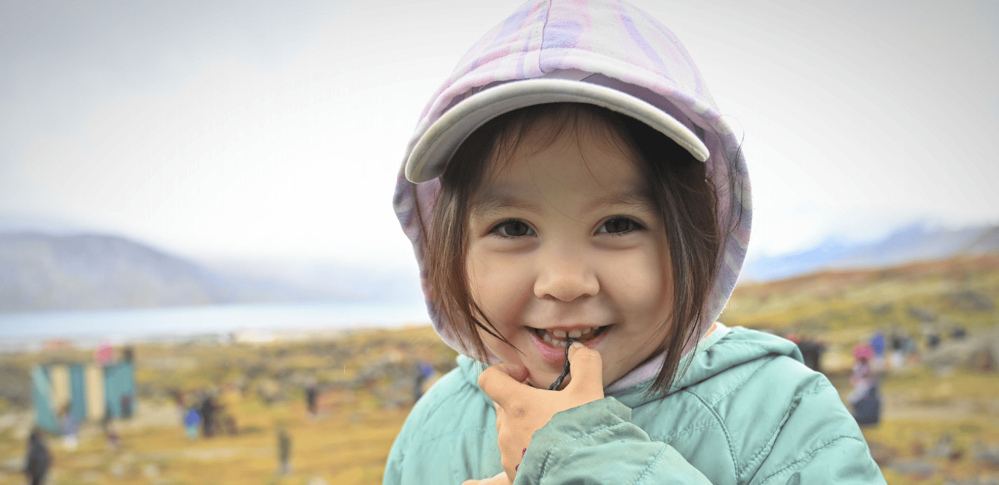 Un·e jeune portant une casquette et une veste se tient à l'extérieur, souriant à l'objectif, une main près de la bouche, avec un paysage naturel flou à l'arrière-plan.