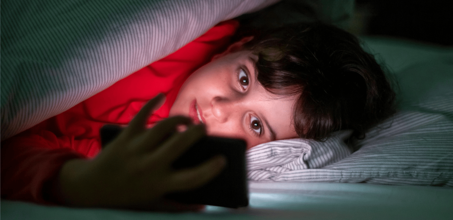 Un·e jeune allongé·e dans un lit sous une couverture, dans une lumière tamisée, regardant un téléphone intelligent qui éclaire son visage.