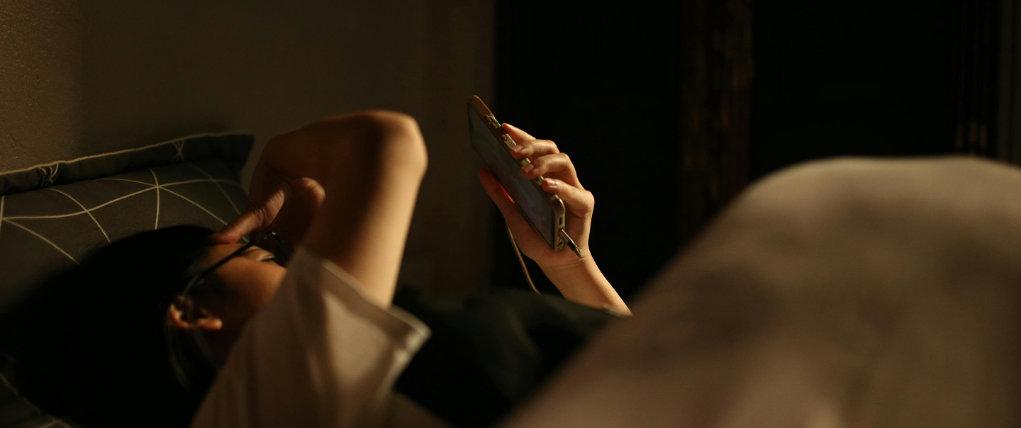 Une personne est allongée sur un lit dans un éclairage chaud et tamisé, tenant un téléphone intelligent au-dessus de son visage, l'autre bras reposant au-dessus de sa tête.