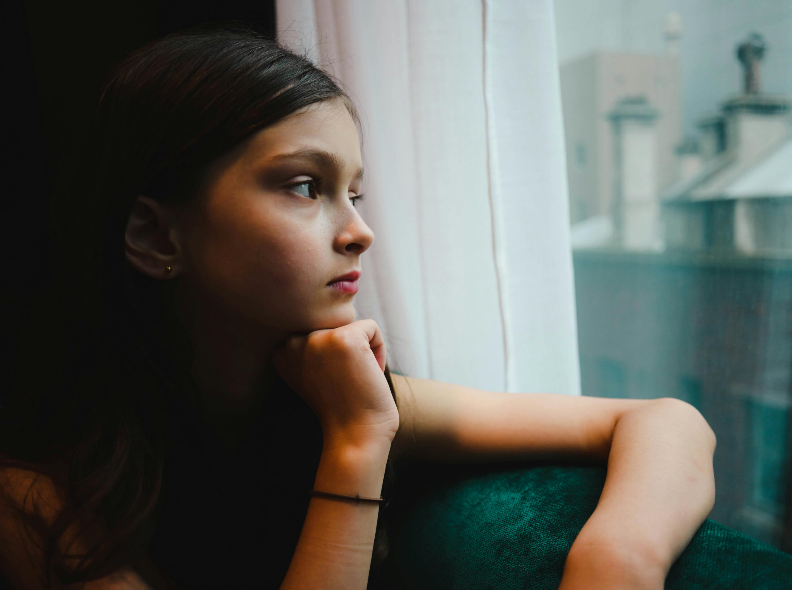 Une personne assise à l'intérieur près d'une fenêtre, le menton appuyé sur la main, le regard tourné vers l'extérieur.